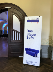 Das Foto zeigt einen Teil des Eingangsbereichs zum Kongresssaal. Vor der Flügeltür steht ein Aufsteller. Darauf ist ein blaues Sofa abgebildet, darüber der Titel 