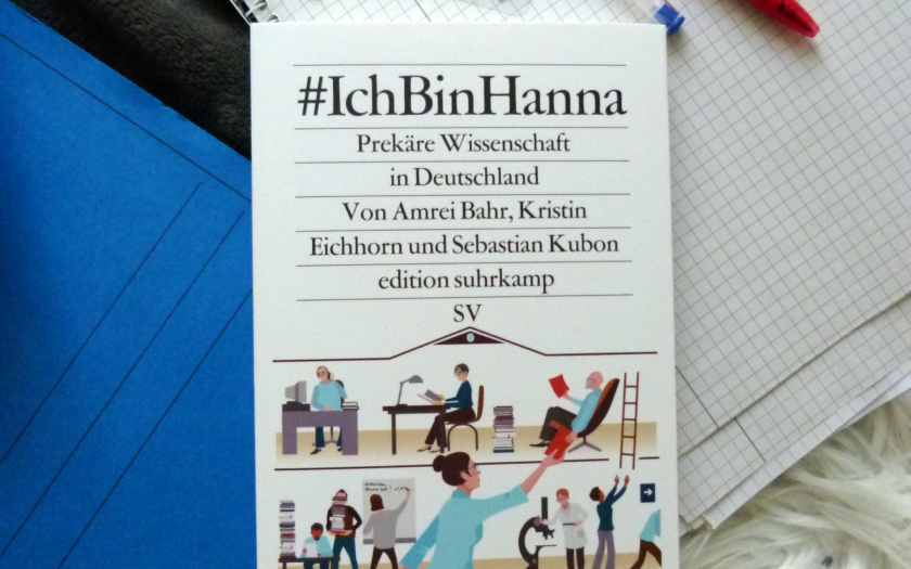 Aus der Vogelperspektive fotografiert: Das Buch liegt zentriert. Oben rechts liegt das Buch auf einem College-Block, darauf liegen bunte Post-Its sowie ein blauer und ein roter Kugelschreiber. Unten links liegt das Buch auf einem blauen Pappschnell-hefter.