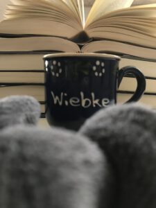 Eine blaue Tasse mit dem Schriftzug "Wiebke" steht vor einem Stapel aufgeschlagener Bücher. Im Vordergrund sind Falten einer grauen Decke zu sehen.