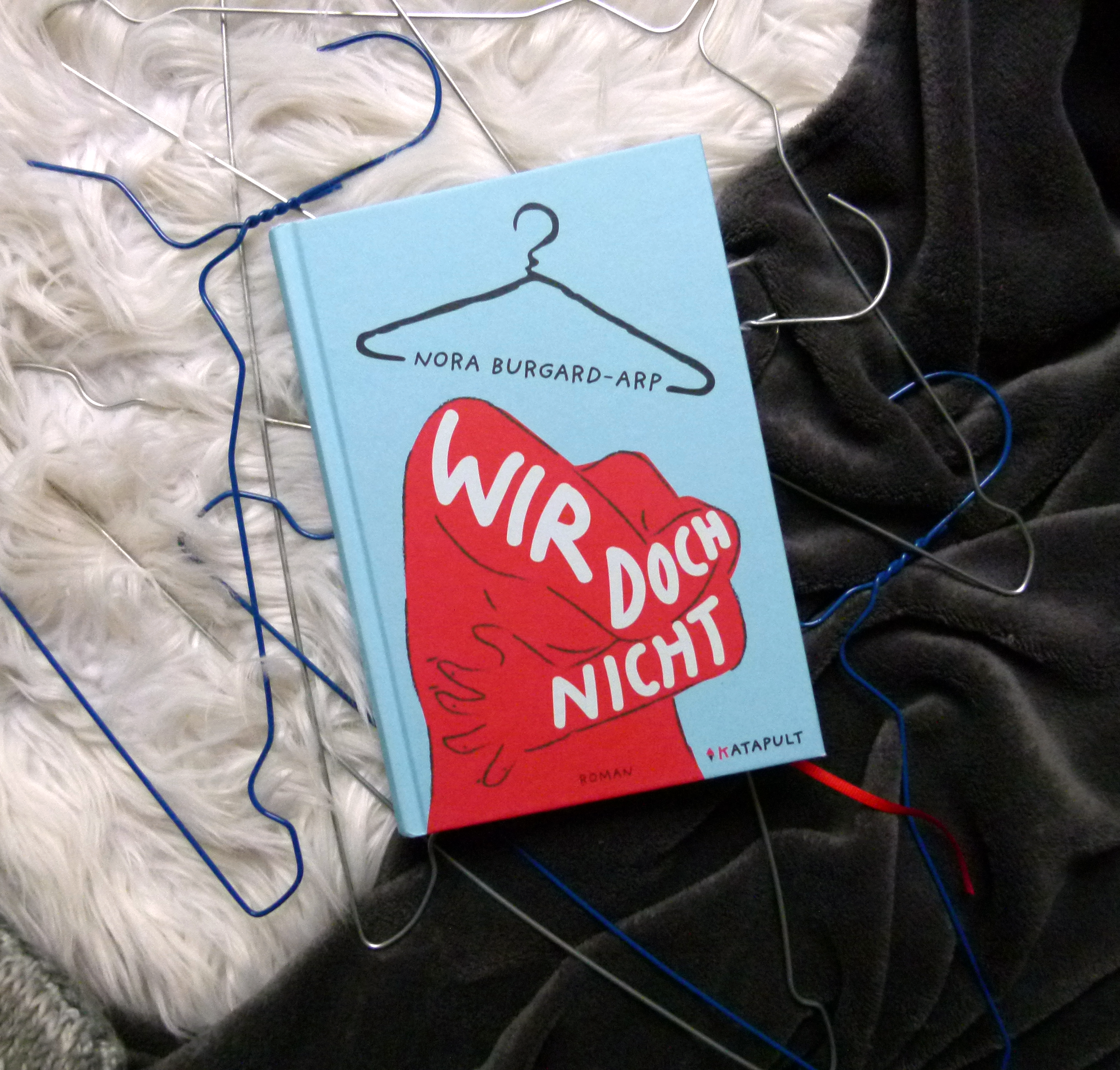 Unter dem Buch liegen zahlreiche Drahtkleiderbügel in unterschiedlichen Farben