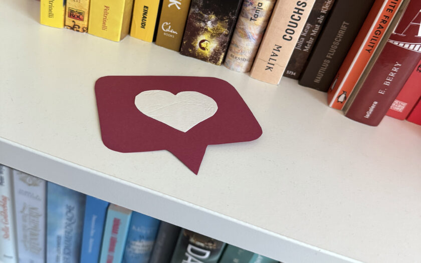 Das Foto zeigt einen Ausschnitt eines nach Farben sortiertes Bücherregal. Auf einem Regalbrett liegt eine rote Sprechblase aus Pappe, auf der ein weißes Herz klebt. Das soll die Benachrichtigung des Instagram-Likes nachahmen.