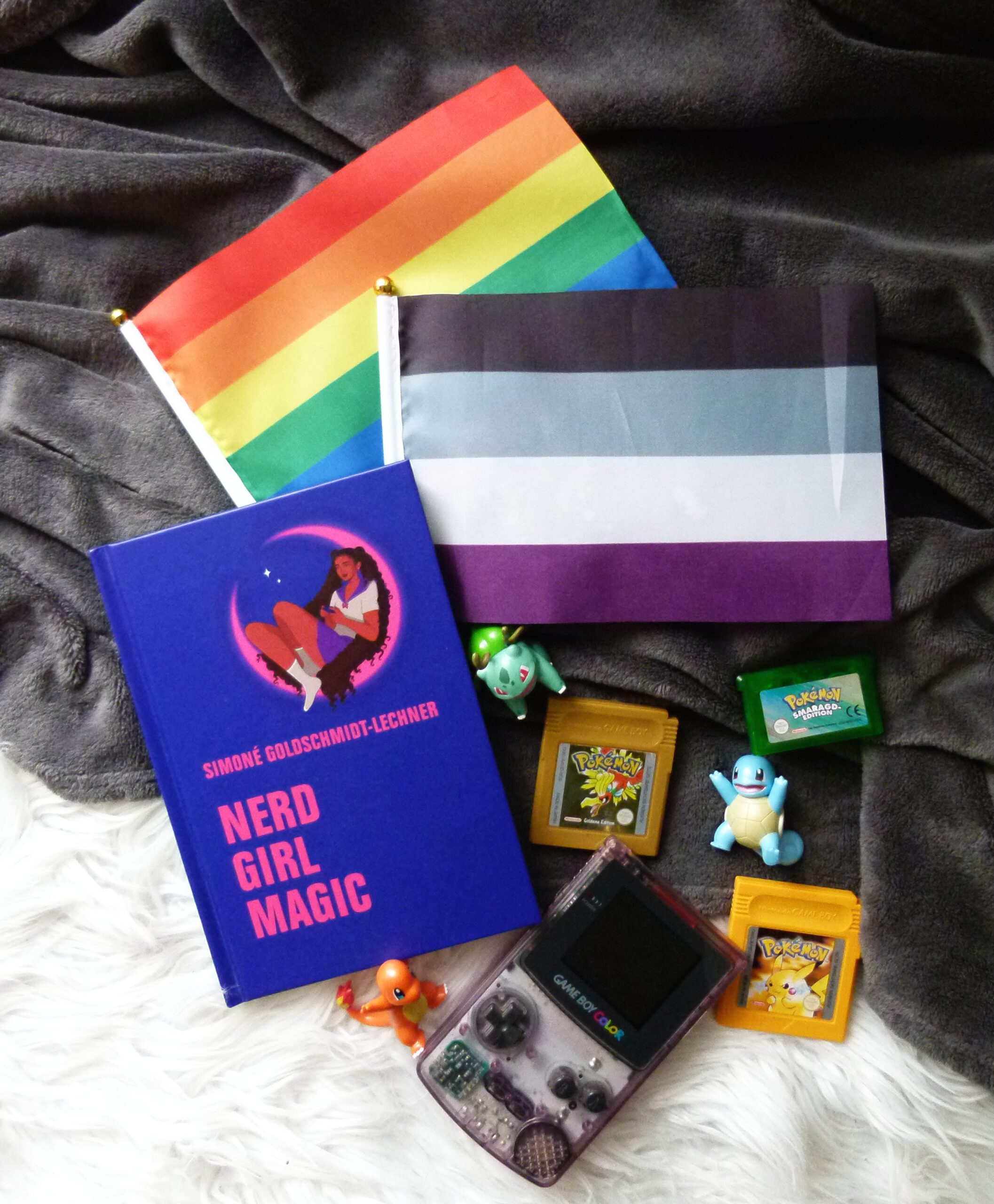 Das Foto zeigt das Sachbuch Nerd Girl Magic von Simoné Goldschmidt-Lechner umgeben von Pokémon-Figuren, Pokémon-Spielkartuschen, einem Gameboy Color und einer kleinen Regenbogen- und Ace-Flagge.