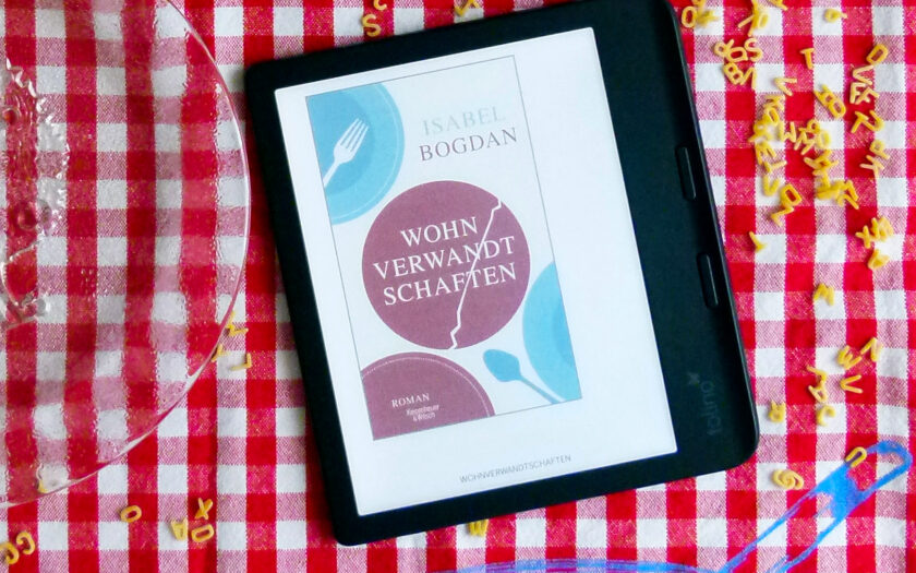 Das Bild zeigt einen rot-weißen karierte Tischdecke, auf der ein E-Book-Reader liegt. Auf der Tischdecke ist zusätzlich eine blaue Skizze einer Schüssel mit einem Löffel gezeichnet. Auf dem Reader ist das Cover des Romans „Wohnverwandtschaften“ von Isabel Bogdan zu sehen. Auf der Tischdecke verstreut liegen Buchstabennudeln. Ein Teil eines leeren Tellers mit Apfelbaummotiv und ein weißer Unterteller mit rot-goldenen Verzierungen sowie ein türkisches Teeglas sind ebenfalls zu sehen.