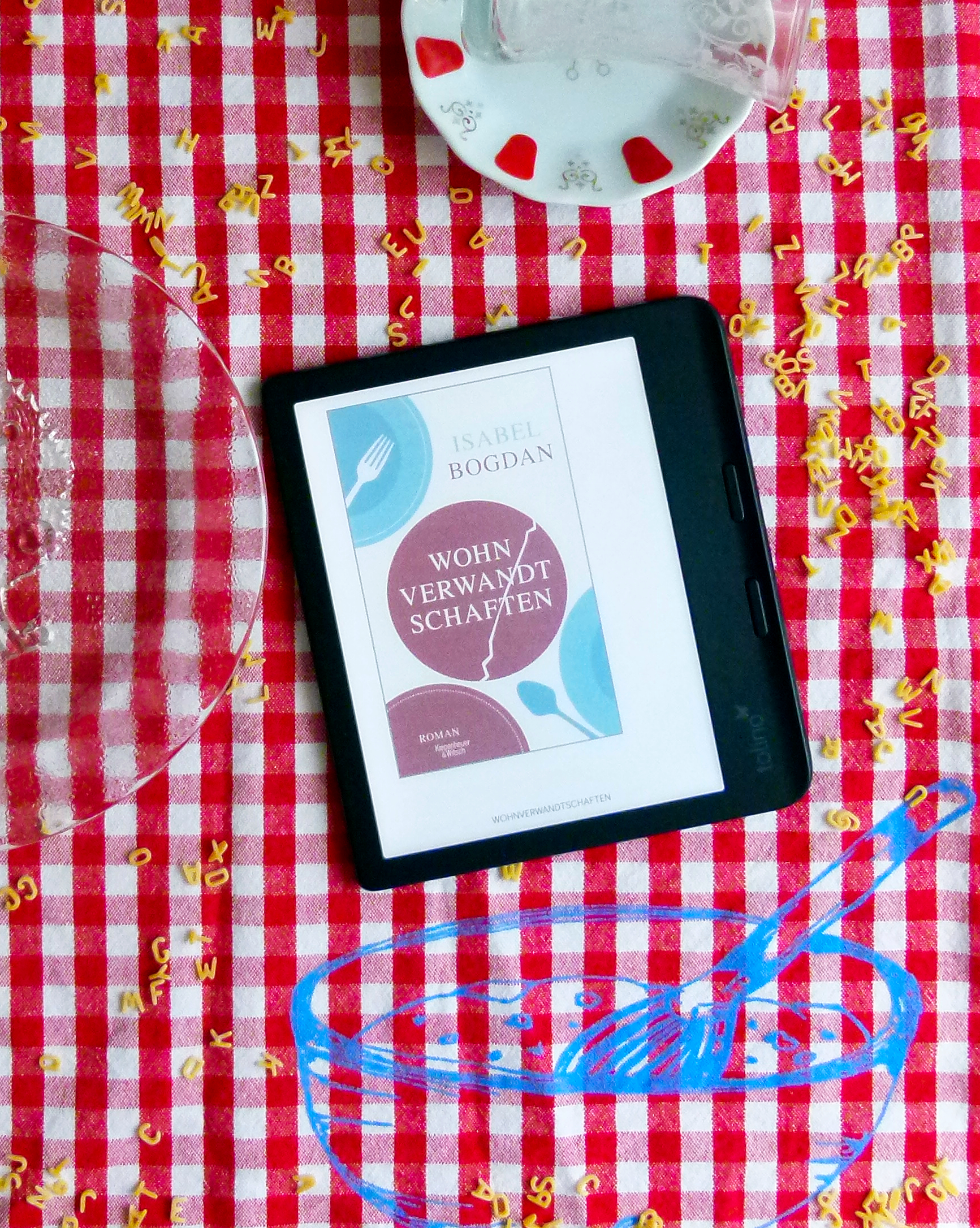Das Bild zeigt einen rot-weißen karierte Tischdecke, auf der ein E-Book-Reader liegt. Auf der Tischdecke ist zusätzlich eine blaue Skizze einer Schüssel mit einem Löffel gezeichnet. Auf dem Reader ist das Cover des Romans „Wohnverwandtschaften“ von Isabel Bogdan zu sehen. Auf der Tischdecke verstreut liegen Buchstabennudeln. Ein Teil eines leeren Tellers mit Apfelbaummotiv und ein weißer Unterteller mit rot-goldenen Verzierungen sowie ein türkisches Teeglas sind ebenfalls zu sehen.