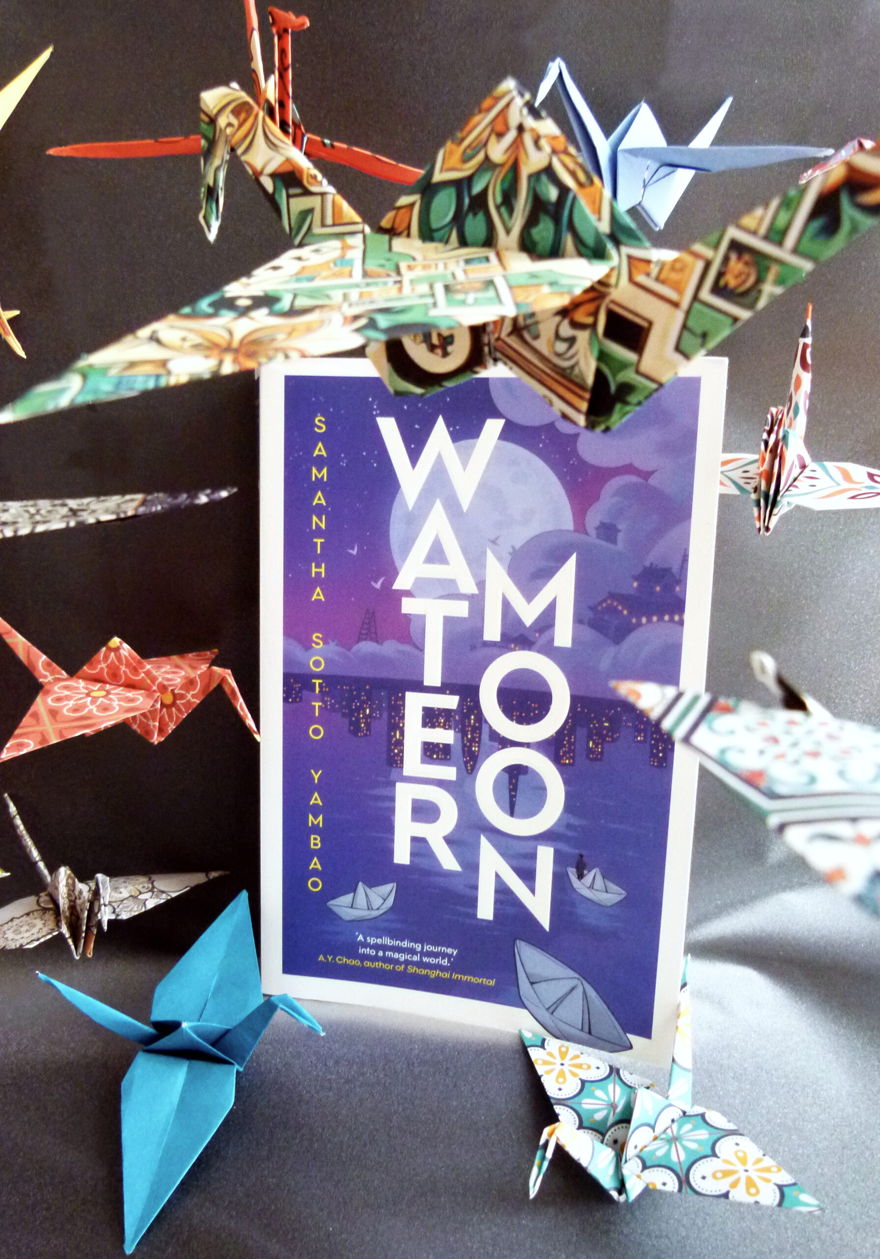 Das Foto zeigt den Roman Water Moon von Samantha Sotto Yambao auf einem dunklen Hintergrund. Um das Buch herum schweben Origami-Kraniche in bunten Farben.