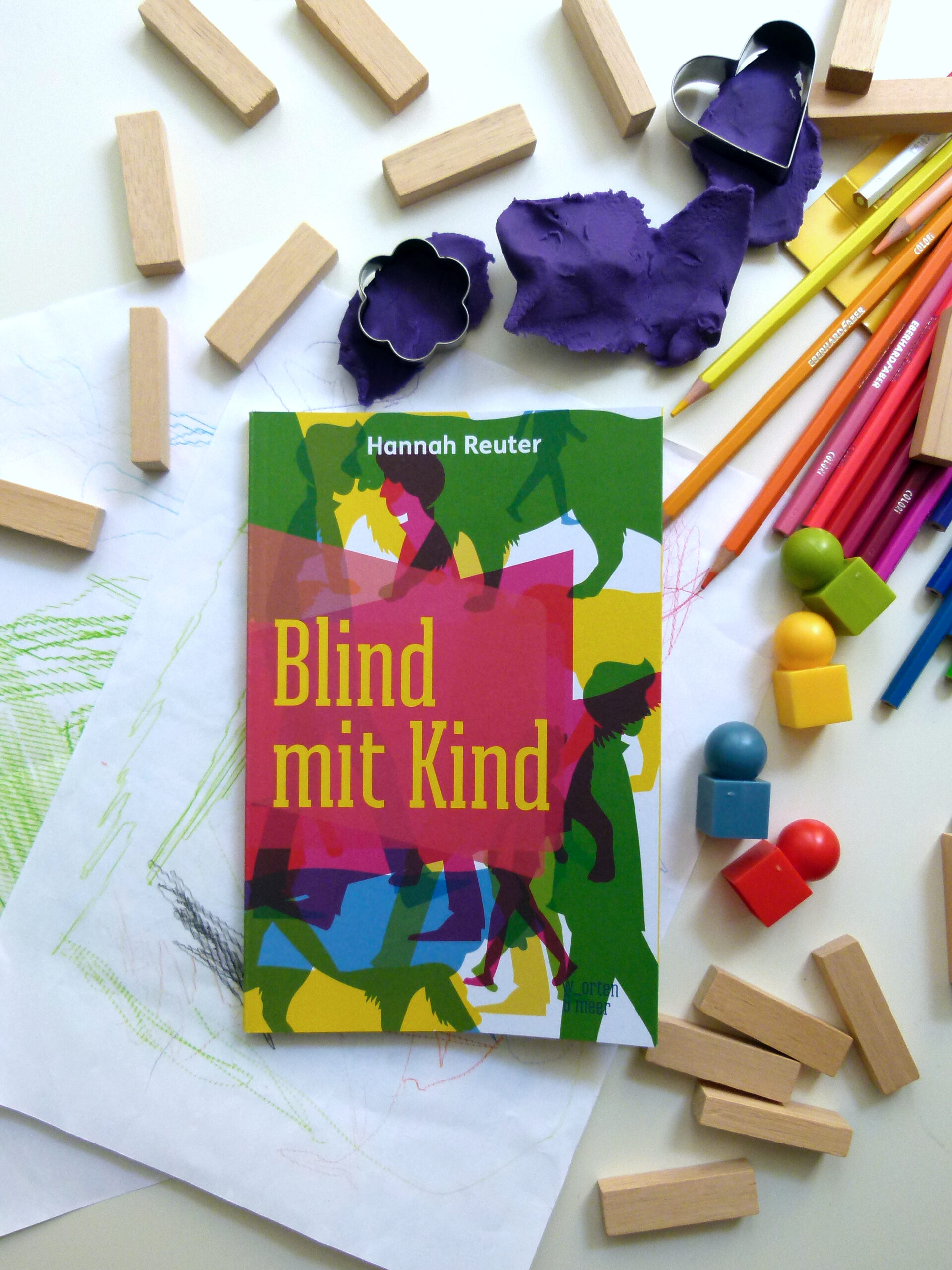 Das Buch liegt zwischen jeder Menge Kinderspielzeug und gemalten Bildern: Knete, Buntstifte, Bauklötze und Spielfiguren liegen überall verteilt.