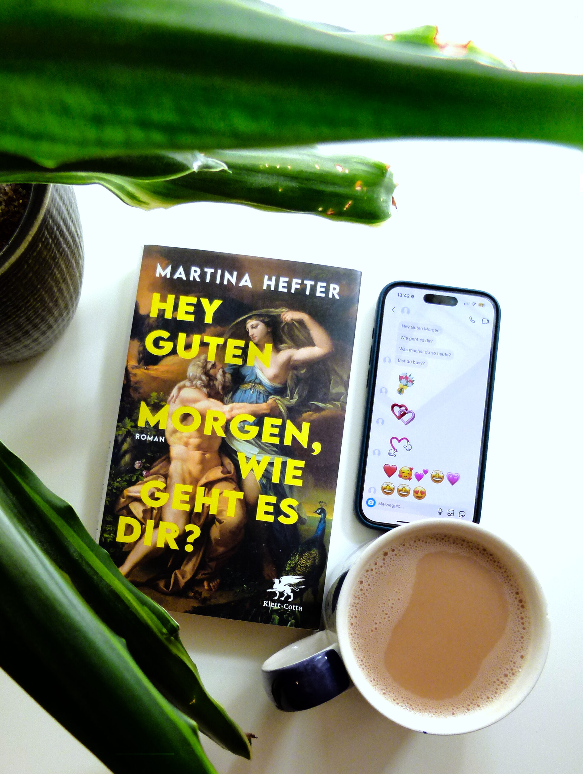 Neben dem Buch liegt ein Handy, dessen Display ua viele Herz-Emojis zeigt. Rechts daneben steht eine Tasse Tee und links daneben steht eine Pflanze, deren Blätter in das Foto hineinreichen.