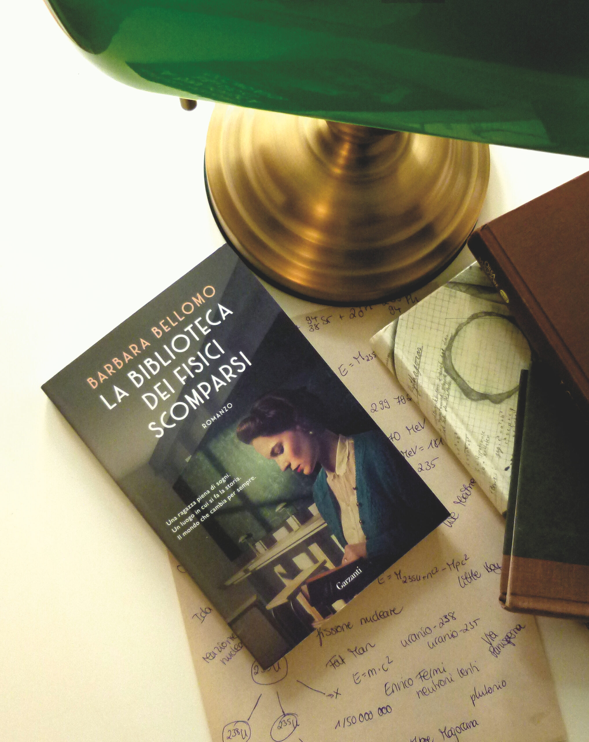 Das Buch liegt auf einem Zettel, auf dem verschiedene Notizen (Formeln, Zahlen etc) zu erkennen sind. Rechts daneben steht ein kleiner Bücherstapel und ein grüner Lampenschirm und -fuß ragen ins Bild hinein. 