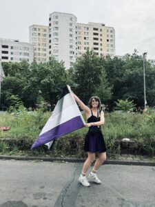 Junge Frau läuft mit einer im Wind flatternden Flagge der asexuellen Community über eine Straße und grinst in die Kamera.