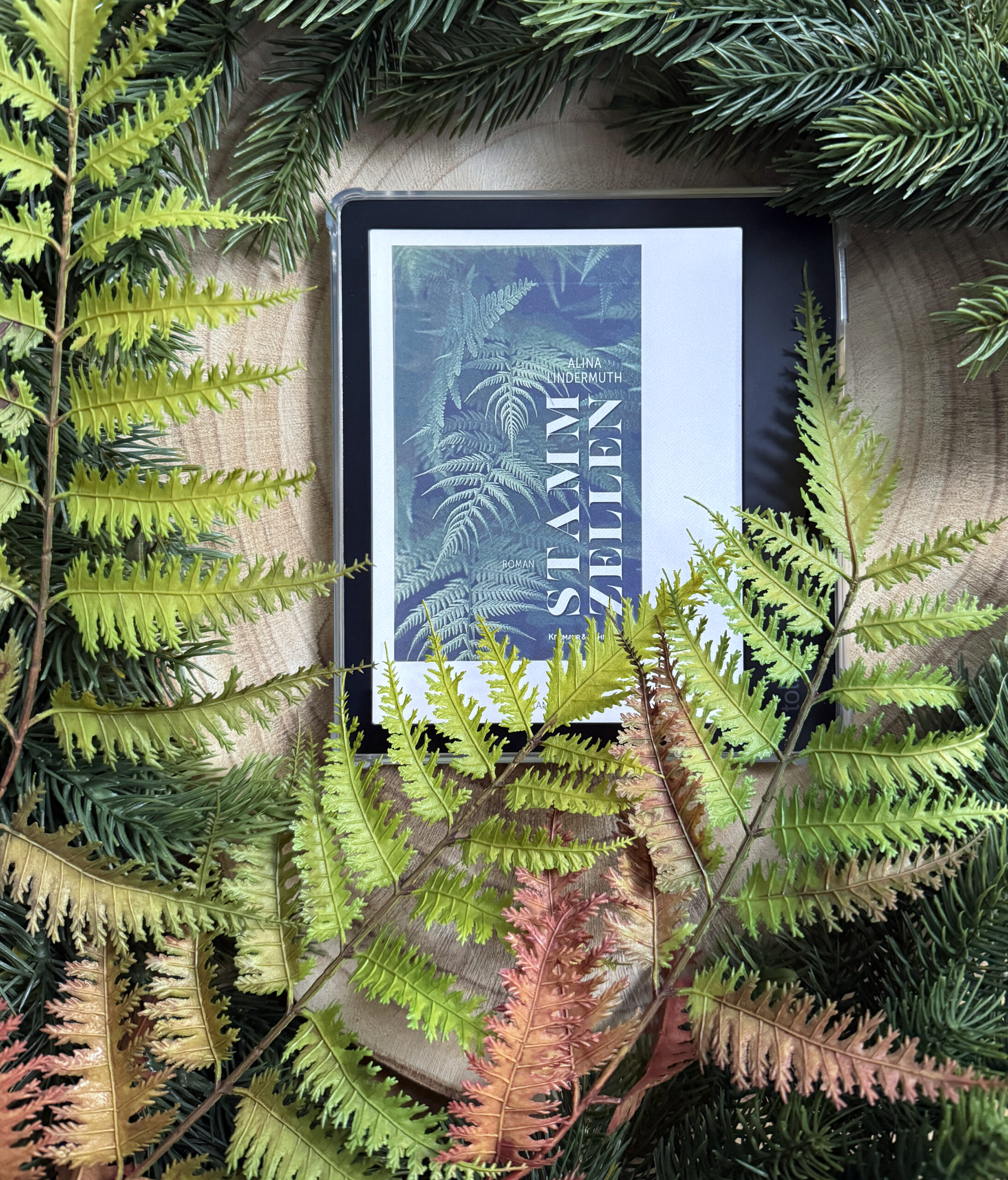 Der eReader mit dem farbigen Cover liegt auf einer Baumscheibe, umgeben von Farnen und Tannenzweigen (aus Plastik).