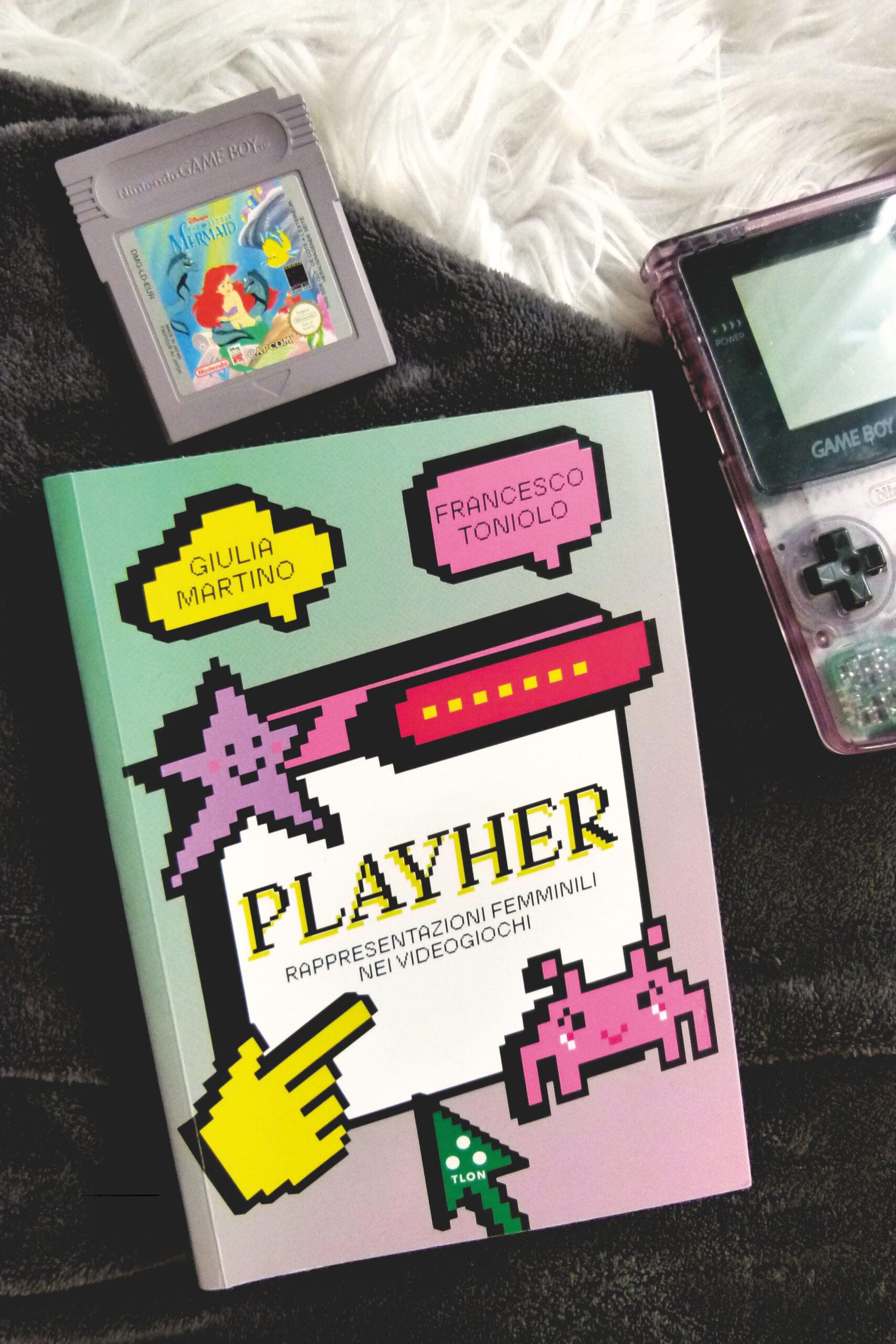 Buch aus der Vogelperspektive fotografiert. Rechts oben liegt ein violett-transparenter GameBoy Color. Links oben liegt die Spielkasette 