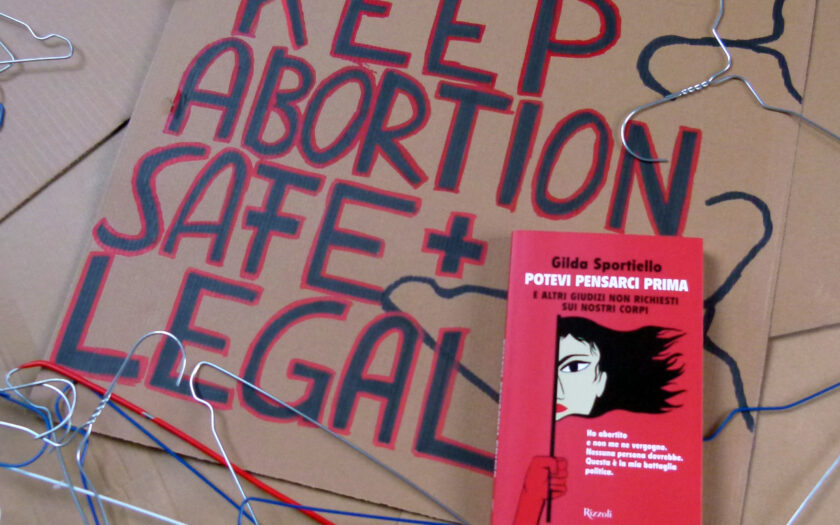 Das Buch liegt auf einem Demo-Plakat auf dem steht: "Keep Abortion Save & Legal". Um das Plakat herum liegen Kleiderbügel aus Metall.