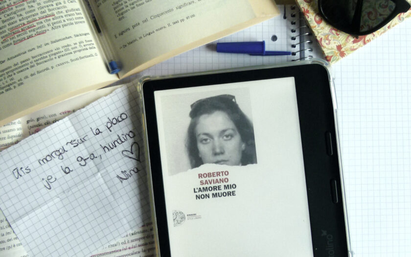 Der eReader, der das Cover des italienischen Romans zeigt liegt auf einem Schreibtisch, umgeben von diversen Gegenständen, wie italienischen Lehrbüchern oder einem Notizzettel auf dem eine Nachricht steht.