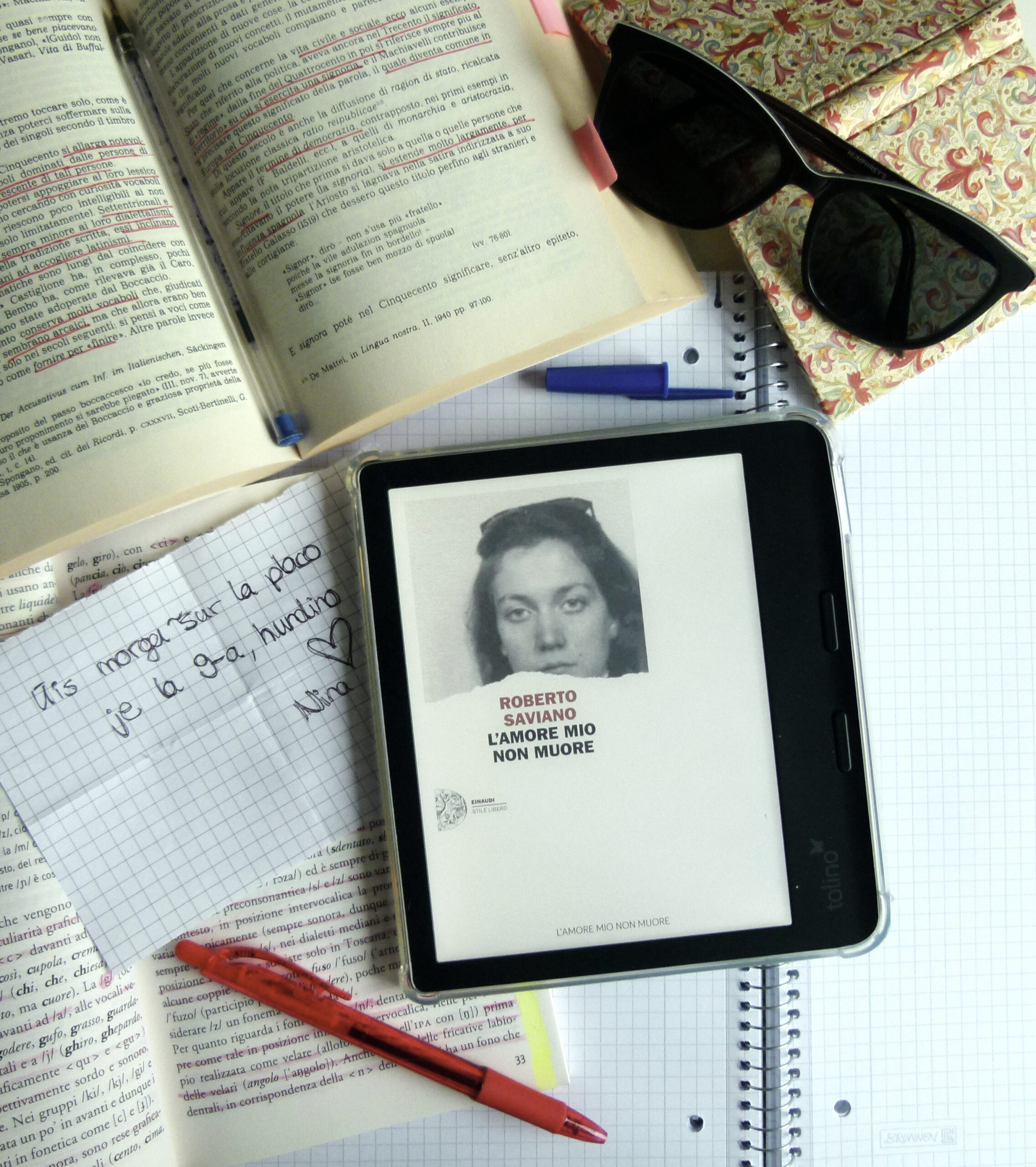 Der eReader, der das Cover des italienischen Romans zeigt liegt auf einem Schreibtisch, umgeben von diversen Gegenständen, wie italienischen Lehrbüchern oder einem Notizzettel auf dem eine Nachricht steht.