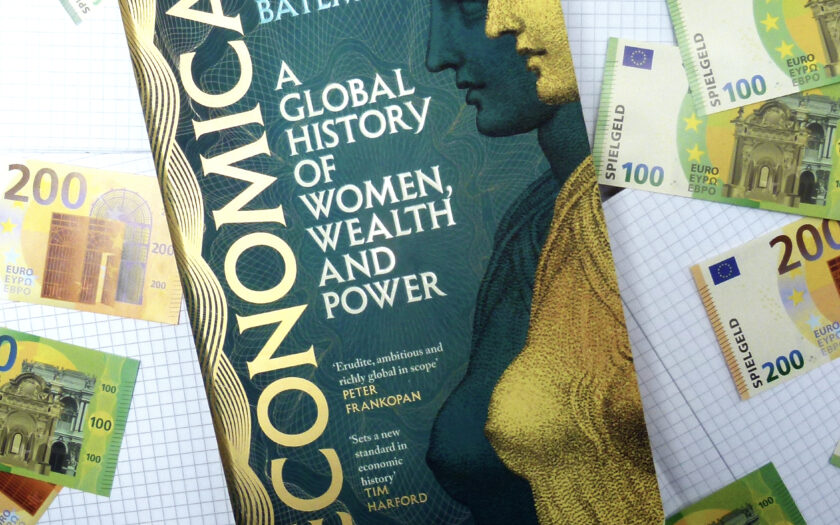 Das Foto zeigt das Buch "Conomica" umgeben von Spielgeld und Spielmünzen, einem Taschenrechner. Das Buch liegt auf kariertem Papier